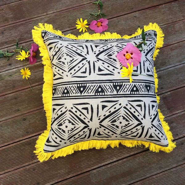 Decorative pillow with black and white pattern and yellow fringes on a wooden surface with flowers.