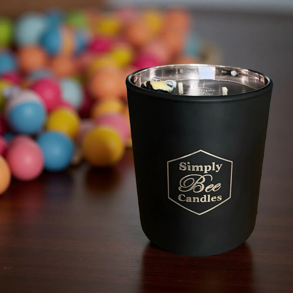 Black candle holder with 'Simply Bee Candles' logo on a wooden surface with colorful balls in the background.