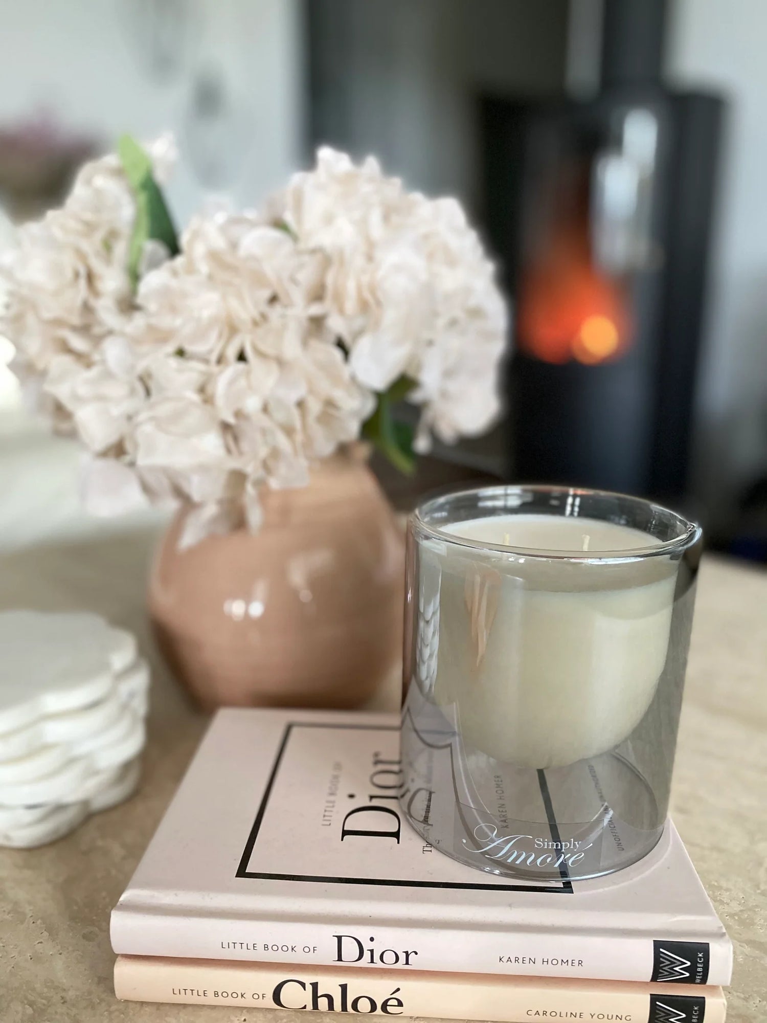 Candle in a glass holder on top of Dior books with flowers in the background