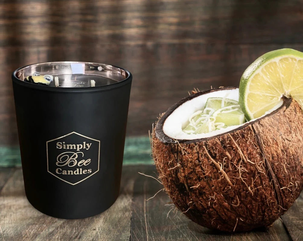 Black candle with 'Simply Bee Candles' branding next to a halved coconut with lime on a wooden surface.