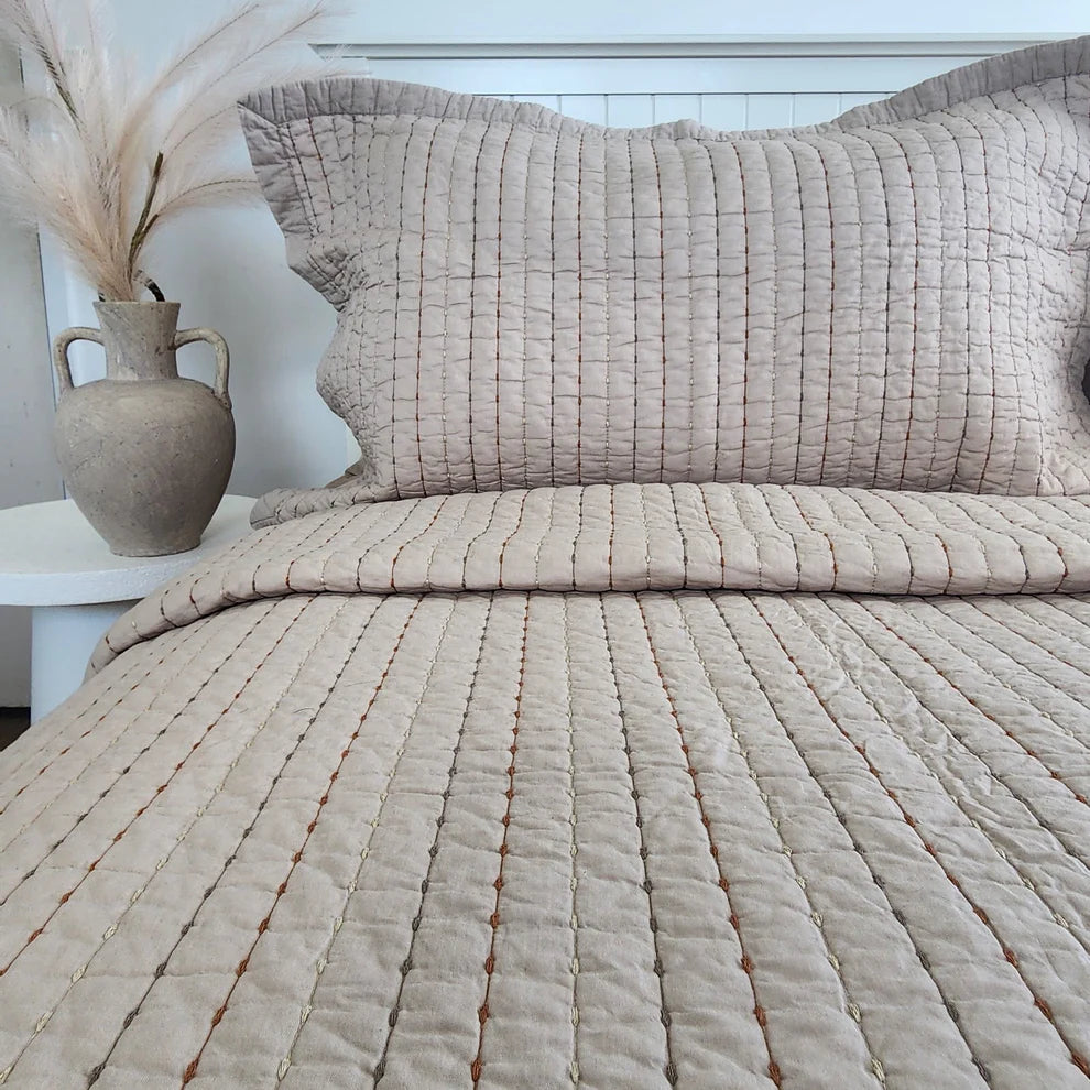Textured beige bedspread and pillows on a bed with a vase and plant in the background.