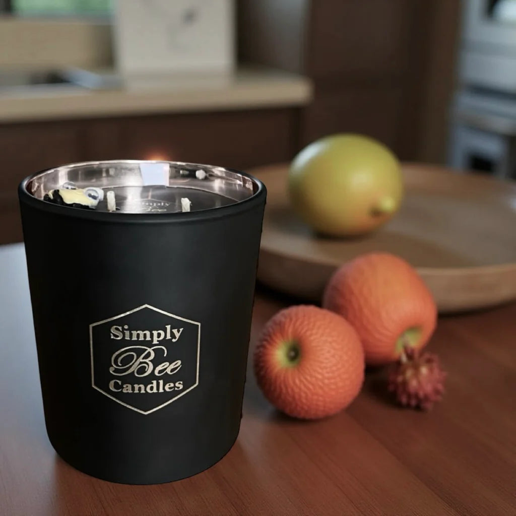 Black candle container with 'Simply Bee Candles' branding on a wooden surface with fruits in the background.