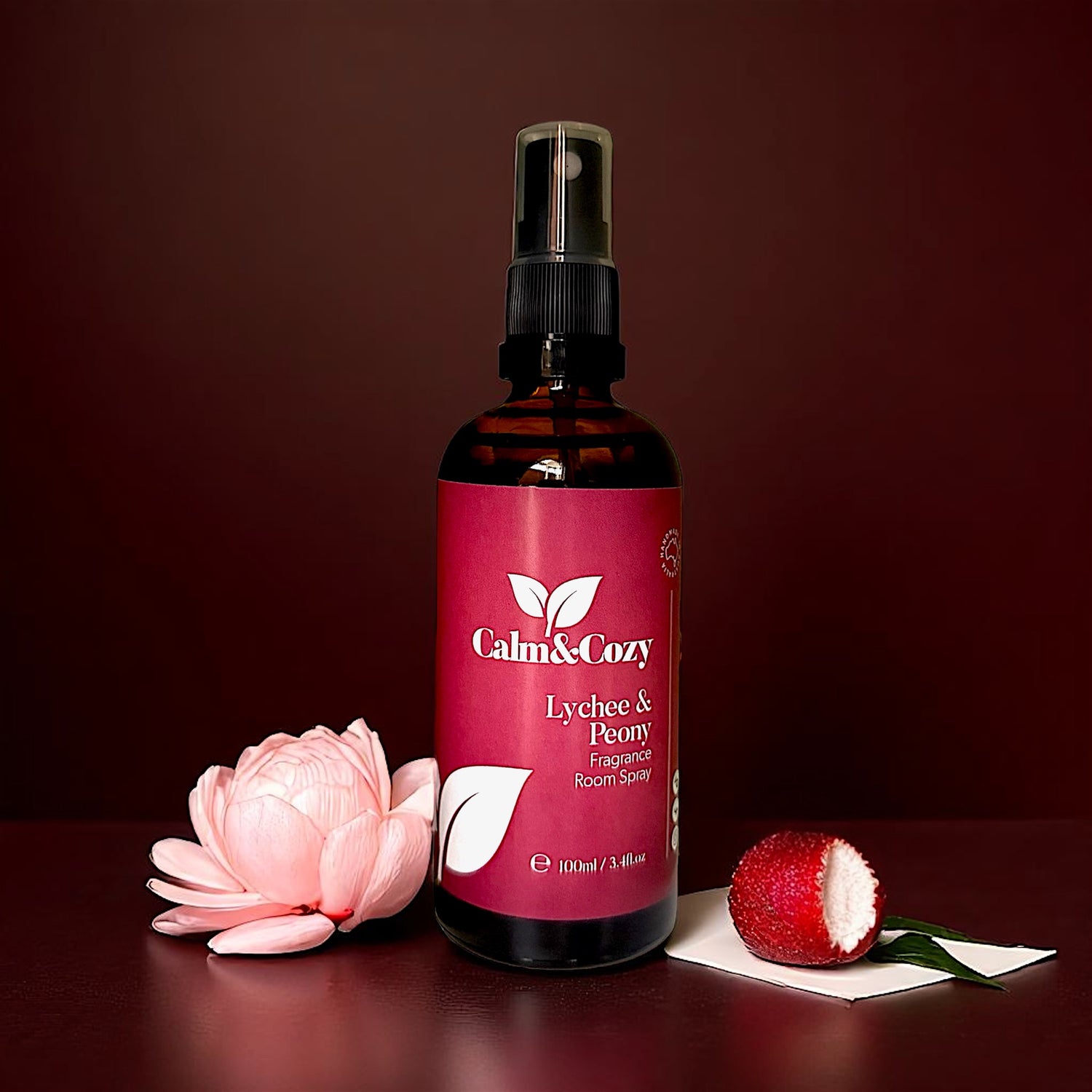 Calm & Cozy lychee & peony room spray bottle with a pink flower and lychee fruit on a dark background