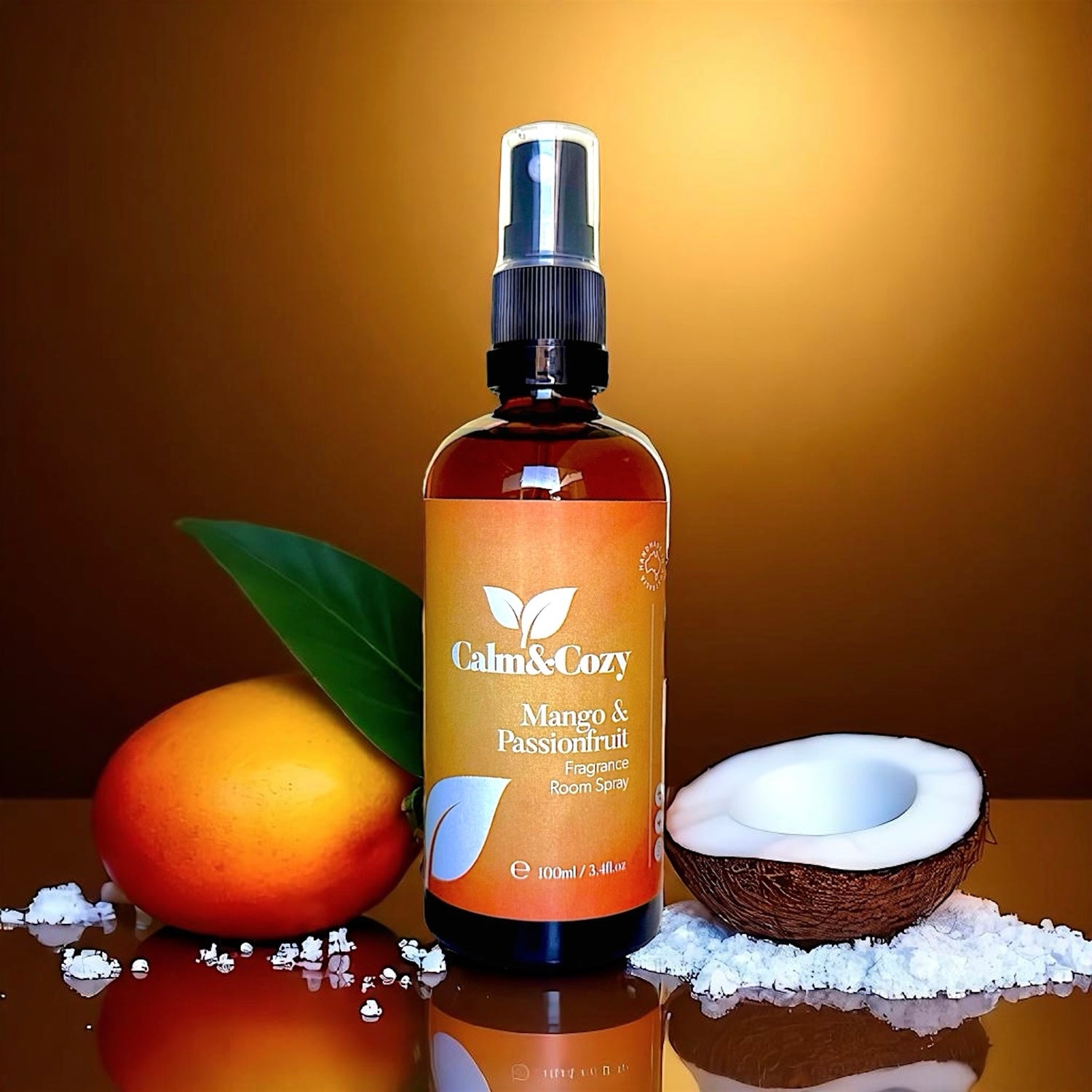 Bottle of Calm & Cozy Mango & Passionfruit room spray with a peach, coconut, and leaf on a reflective surface.