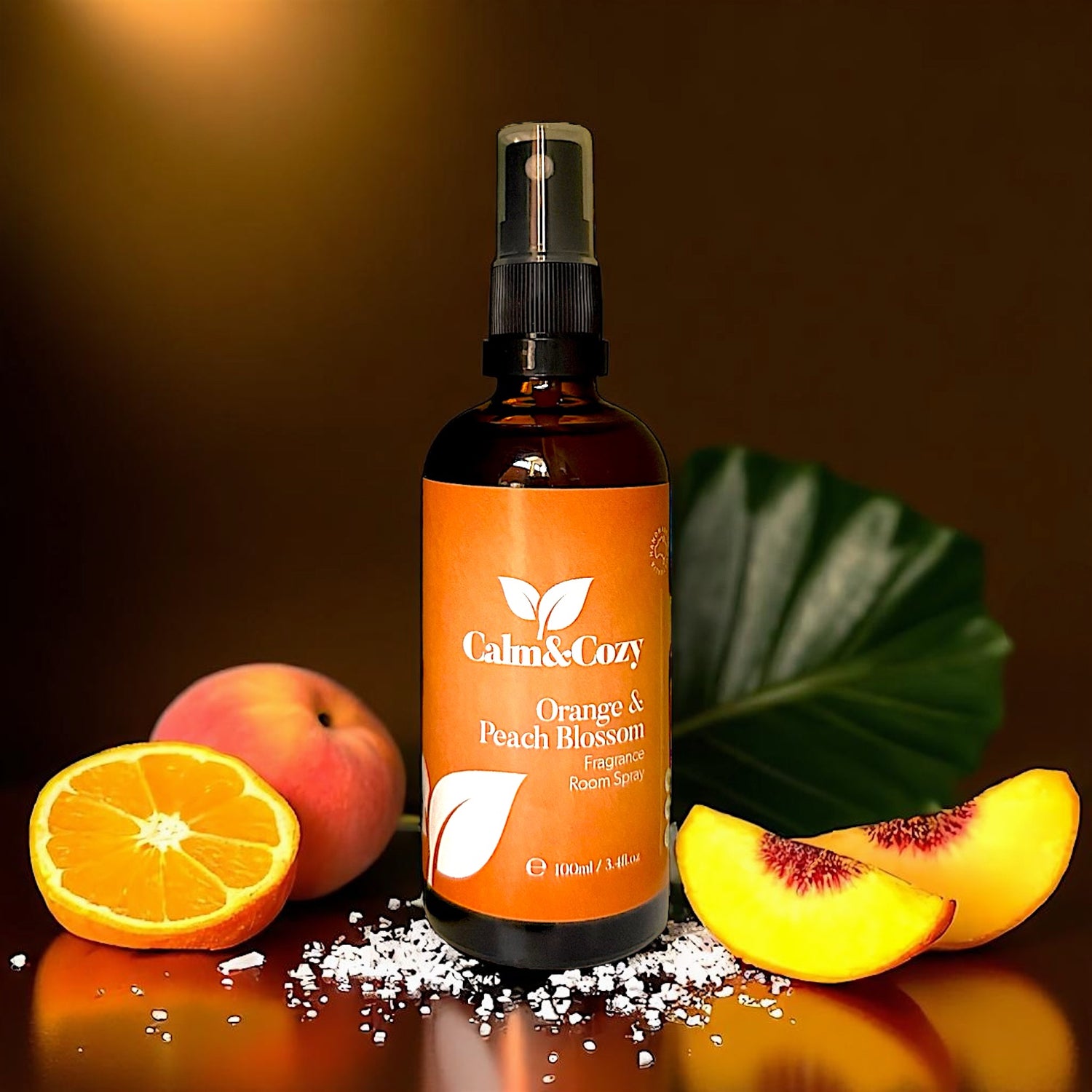 Bottle of Calm & Cozy Orange & Peach Blossom product with fruits and salt on a reflective surface.