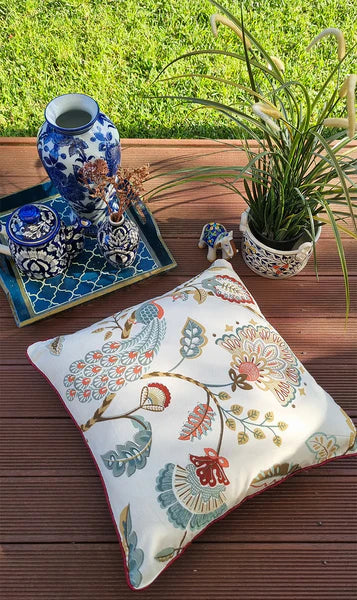 Decorative pillow with floral pattern on a wooden surface with a plant and blue and white vase in the background.
