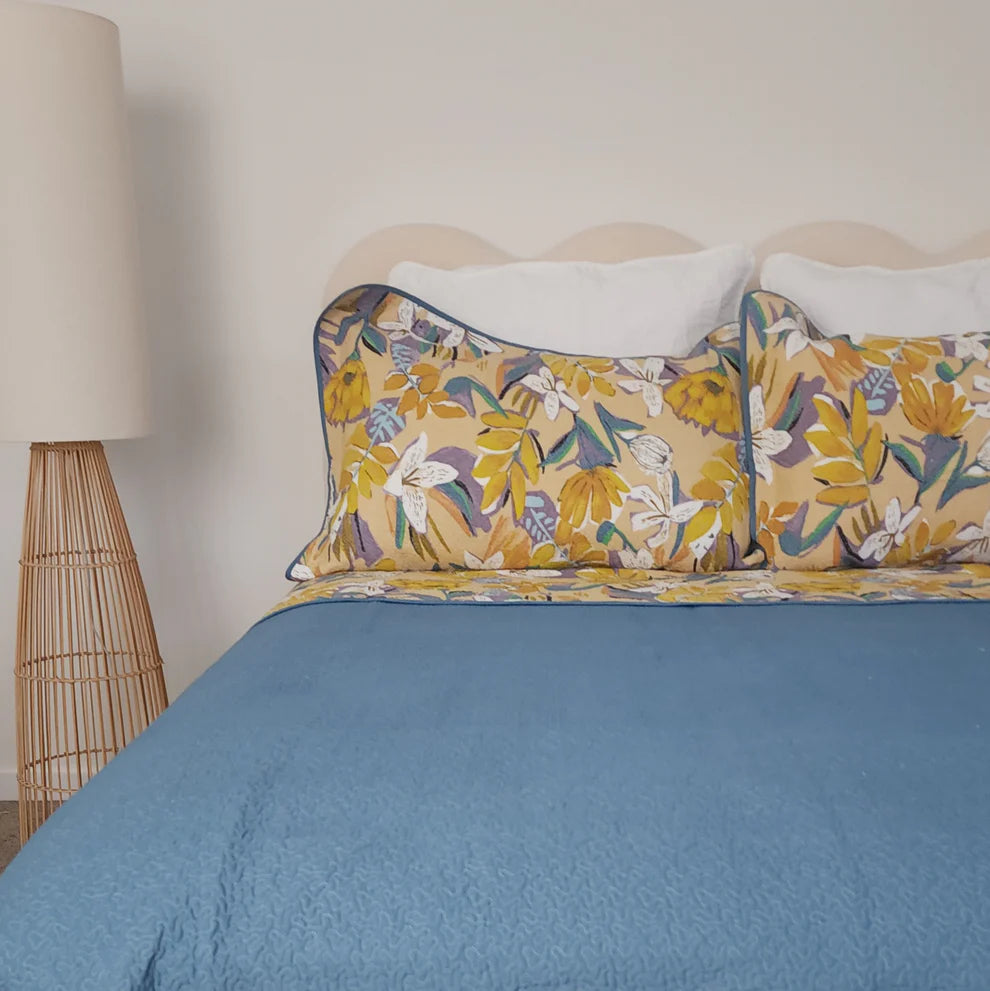 Bed with floral pillows and a blue comforter next to a lamp.