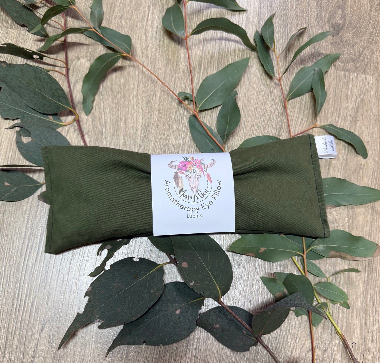 Green therapy eye pillow with a label on a wooden surface with eucalyptus leaves.
