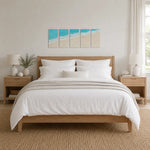A neatly made bed with white bedding and a light wood frame, styled in a bright, modern bedroom. Above the bed hangs a four-panel beach-themed wall art set featuring turquoise ocean waves and beige sand.
