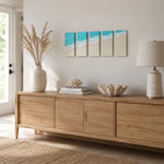 A light natural wood sideboard or credenza in a bright, modern room, styled with a ceramic vase of pampas grass, books, a shell, and a table lamp. Above it hangs a four-panel canvas print depicting a beach and ocean.