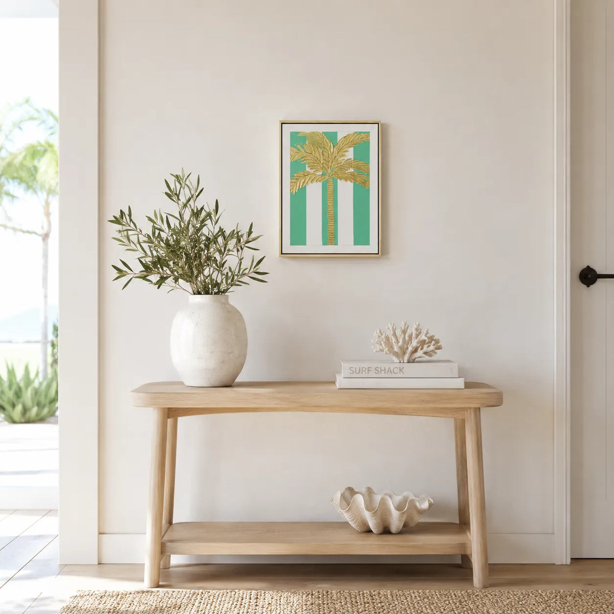 A framed art print featuring a gold palm tree on a teal and white striped background, styled on a light wood console table in a bright, modern room.