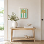 A framed art print featuring a gold palm tree on a teal and white striped background, styled on a light wood console table in a bright, modern room.