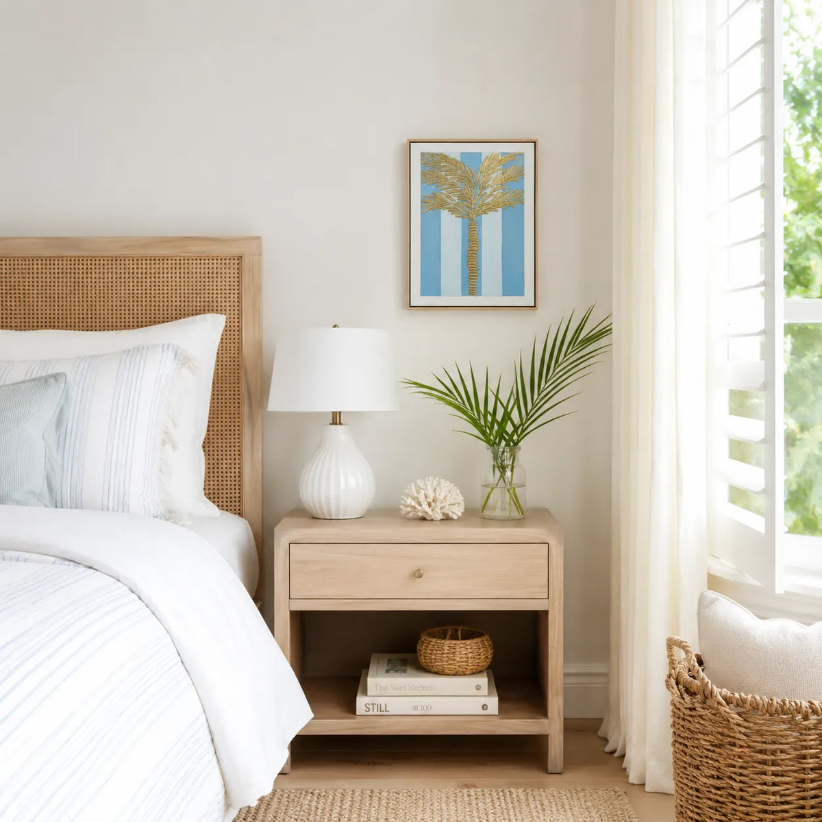 A styled bedroom corner featuring a light wood nightstand with a white ceramic lamp, a small palm plant in a glass vase, and a white shell decor piece. Above the nightstand, a framed art print of a palm tree with gold stripes on a blue background hangs on a beige wall next to a bed with white bedding.