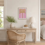 A framed art print featuring a pink and gold palm tree design, styled on a cream wall above a light wood desk with a wicker chair, lamp, and plant.