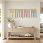 A styled interior scene showing a light wood console table against a white wall, decorated with a white vase of greenery, books, and a woven tray. Above the table are four framed palm tree prints in pastel yellow, green, pink, and blue.