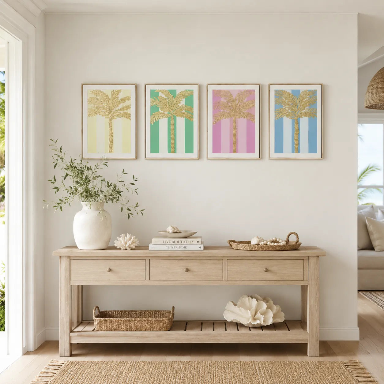 A styled interior scene showing a light wood console table against a white wall, decorated with a white vase of greenery, books, and a woven tray. Above the table are four framed palm tree prints in pastel yellow, green, pink, and blue.