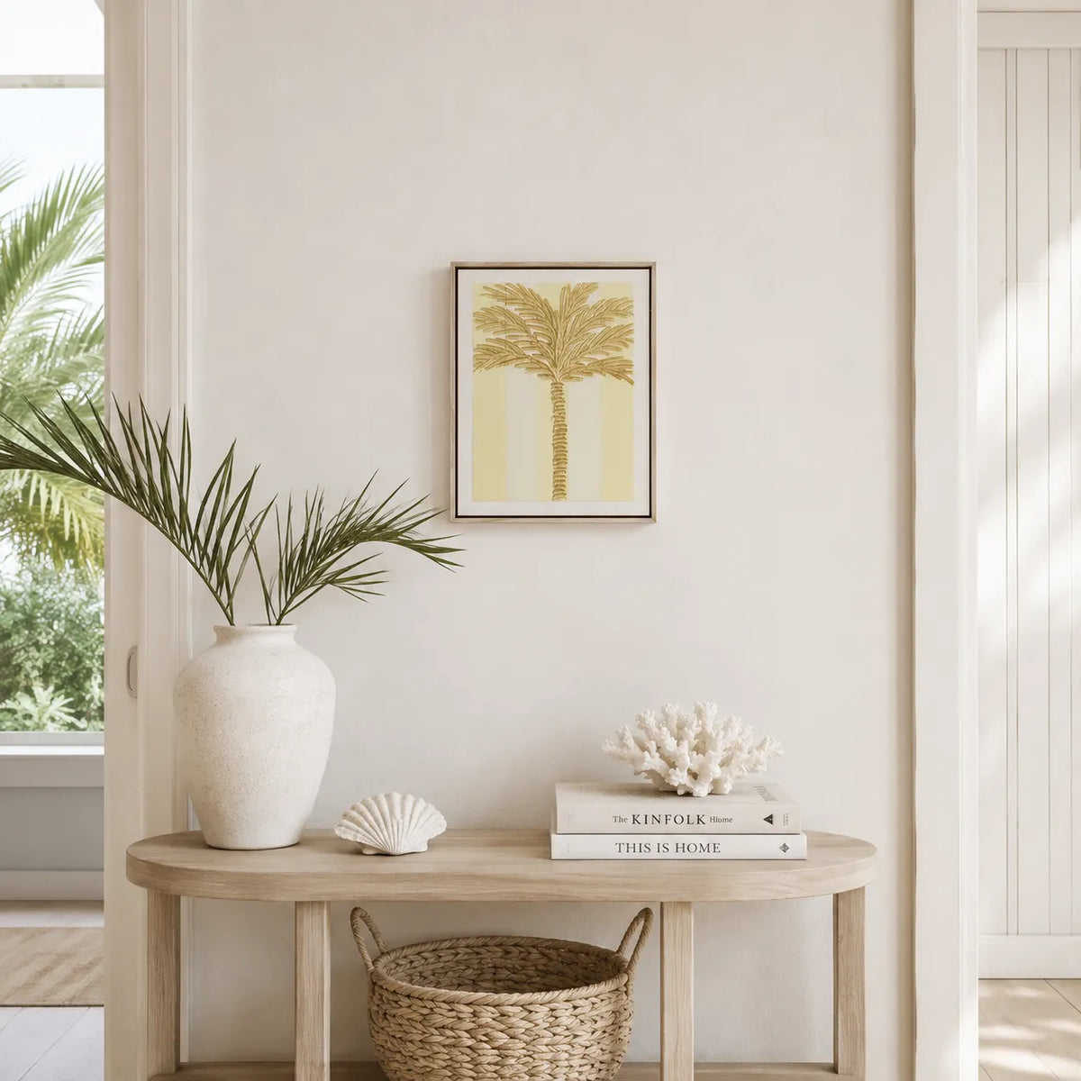 A framed art print of a palm tree in a warm yellow and beige palette, displayed on a cream wall above a light wood console table styled with a white vase, seashells, books, and a woven basket.