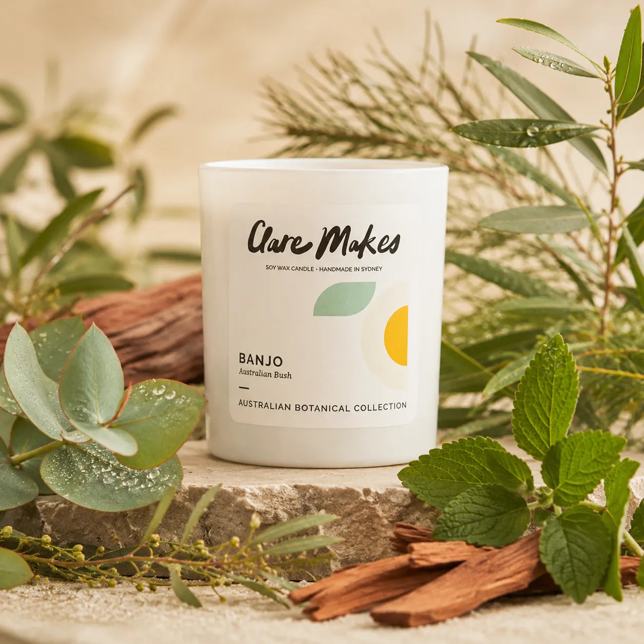 A white glass candle jar labeled 'Care Makes' and 'BANJO Australian Bush' sits on a stone surface surrounded by green leaves, cinnamon sticks, and soft natural light.