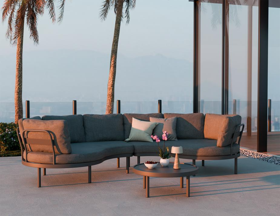 Outdoor patio set with a curved sofa and coffee table on a deck with palm trees and ocean view.