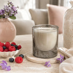 A frosted smoky gray glass candle jar with white wax sits on a round wooden coaster on a light stone table. The setting is a cozy, softly blurred living room scene with a pink vase of purple flowers, a bowl of fresh raspberries, and a neutral-colored sofa in the background.