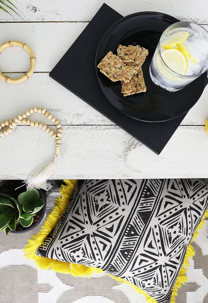 Top image: Black plate with crackers and a glass of water on a white surface. Bottom image: Black and white patterned pillow with yellow fringe on a gray surface.