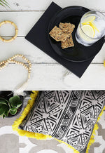 Top image: Black plate with crackers and a glass of water on a white surface. Bottom image: Black and white patterned pillow with yellow fringe on a gray surface.