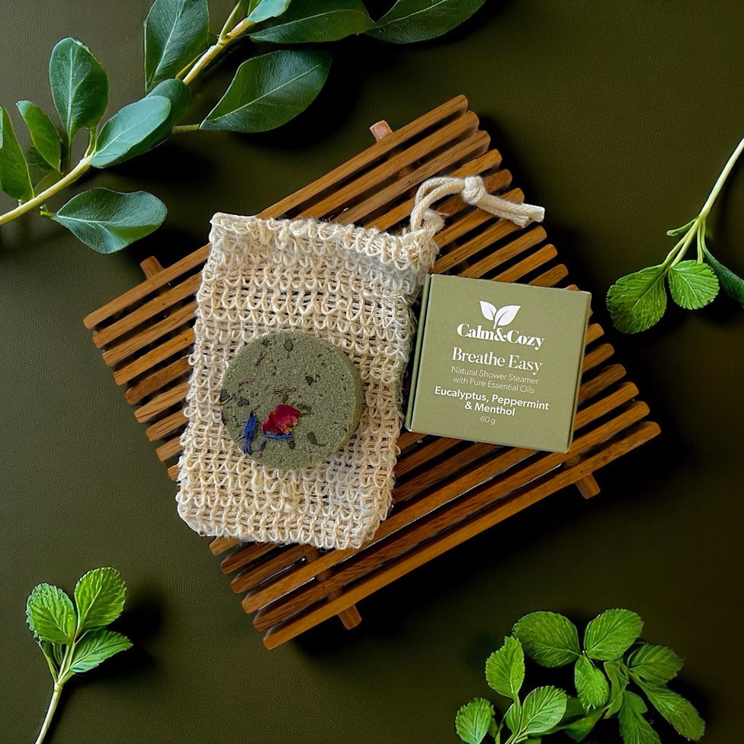Handmade soap in a natural fiber bag with a card on a wooden board, surrounded by green leaves.