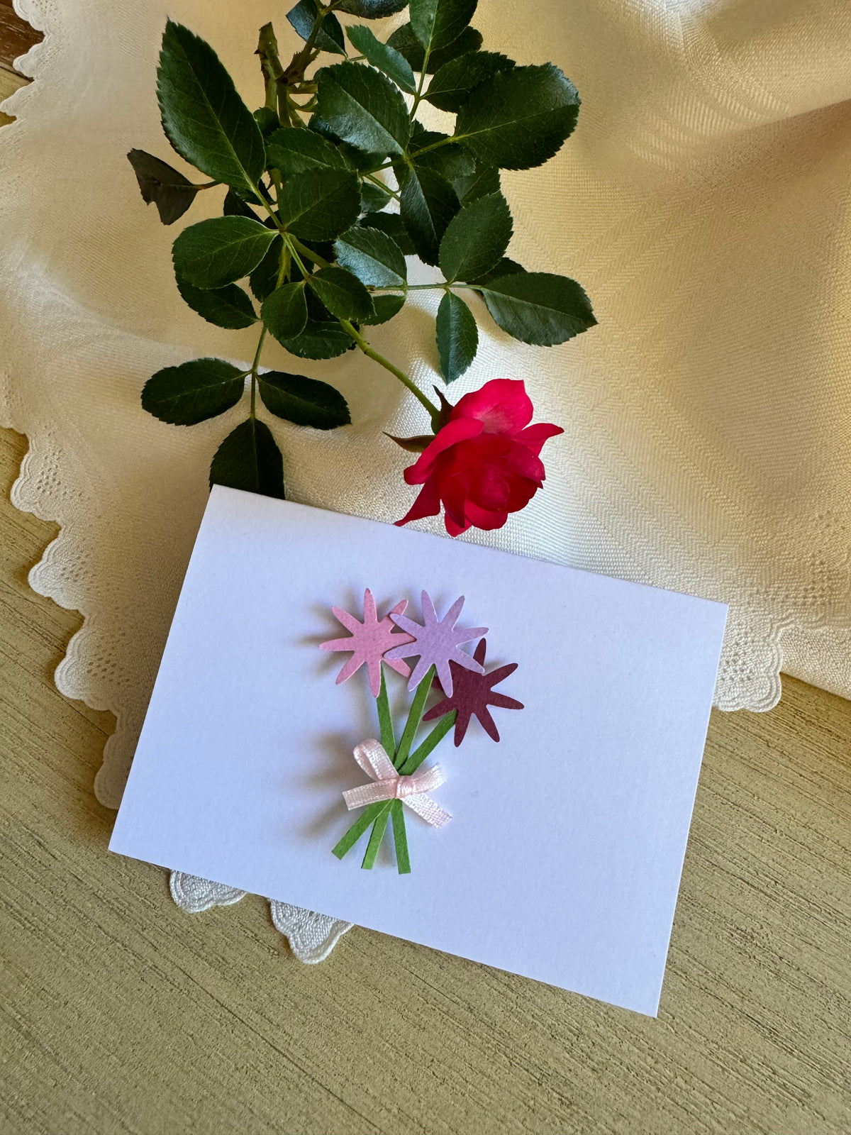Card with paper flowers on a wooden surface with a rose and lace fabric.