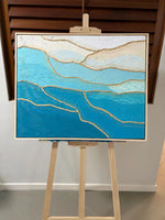 A framed abstract painting with layered blue and white textured waves and gold metallic lines, displayed on a wooden easel in an indoor setting.