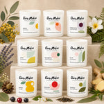 Nine white scented candles from Clare Makes are arranged on beige stone blocks, surrounded by botanical props including green leaves, a white flower, yellow berries, lavender, tea leaves, and ginger root. Each candle features a minimalist label with abstract color shapes and the product name in English and Australian Aboriginal Language.