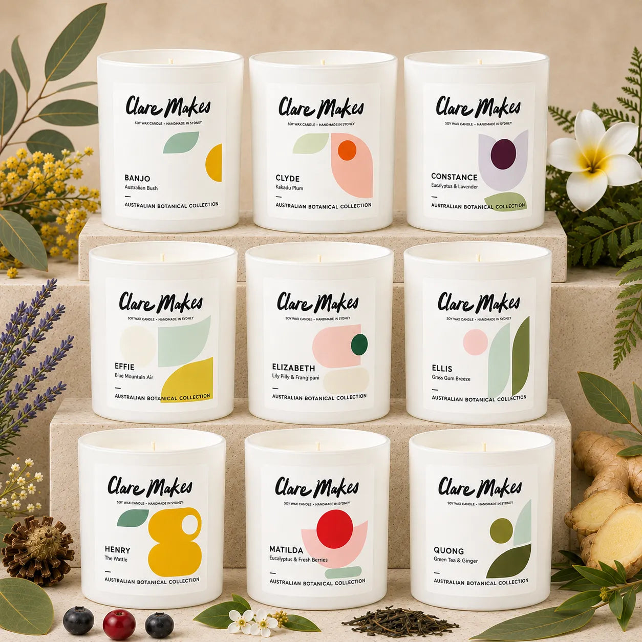 Nine Clare Makes scented candles in white glass jars are arranged in a 3x3 grid on stacked stone blocks, surrounded by botanical props including flowers, leaves, ginger, and berries. Each label features a different botanical design with abstract shapes and scent names such as Banjo, Cyde, Constance, Epifie, Elizabeth, Ellis, Henry, Matilda, and Guong.