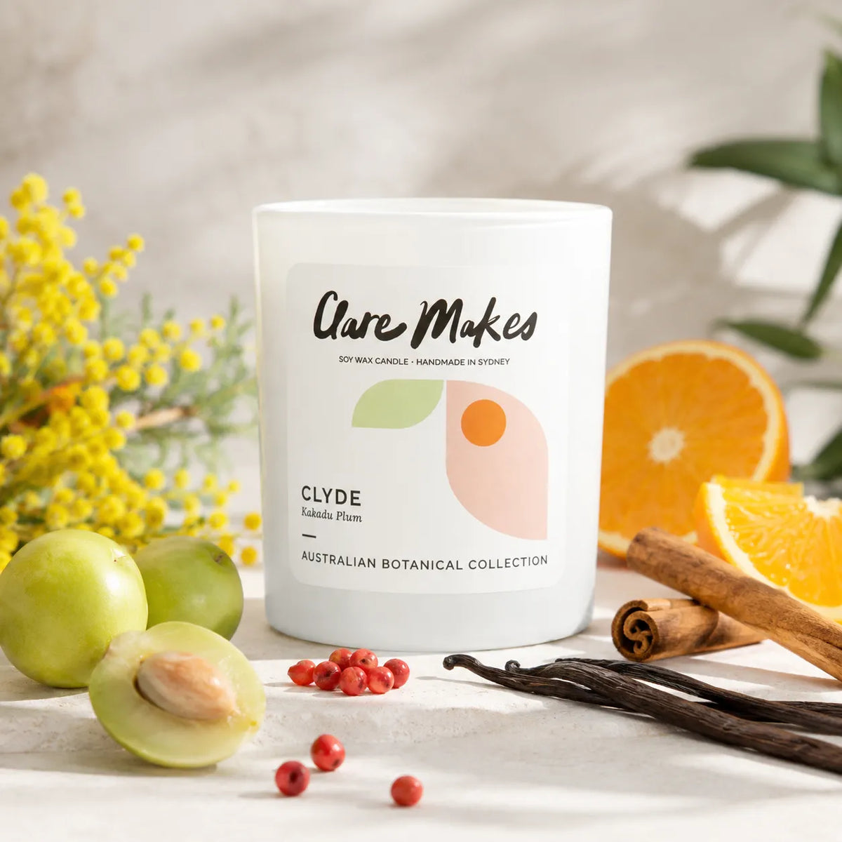 A white soy wax candle jar labeled 'Clyde' by Clare Makes, styled with green plums, yellow flowers, vanilla beans, cinnamon sticks, and orange slices on a light stone surface.