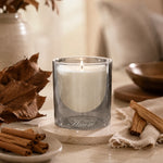 A lit white candle in a clear smoky glass holder sits on a stone coaster on a tabletop, styled with dried leaves, cinnamon sticks, and a neutral fabric in a warm indoor setting.