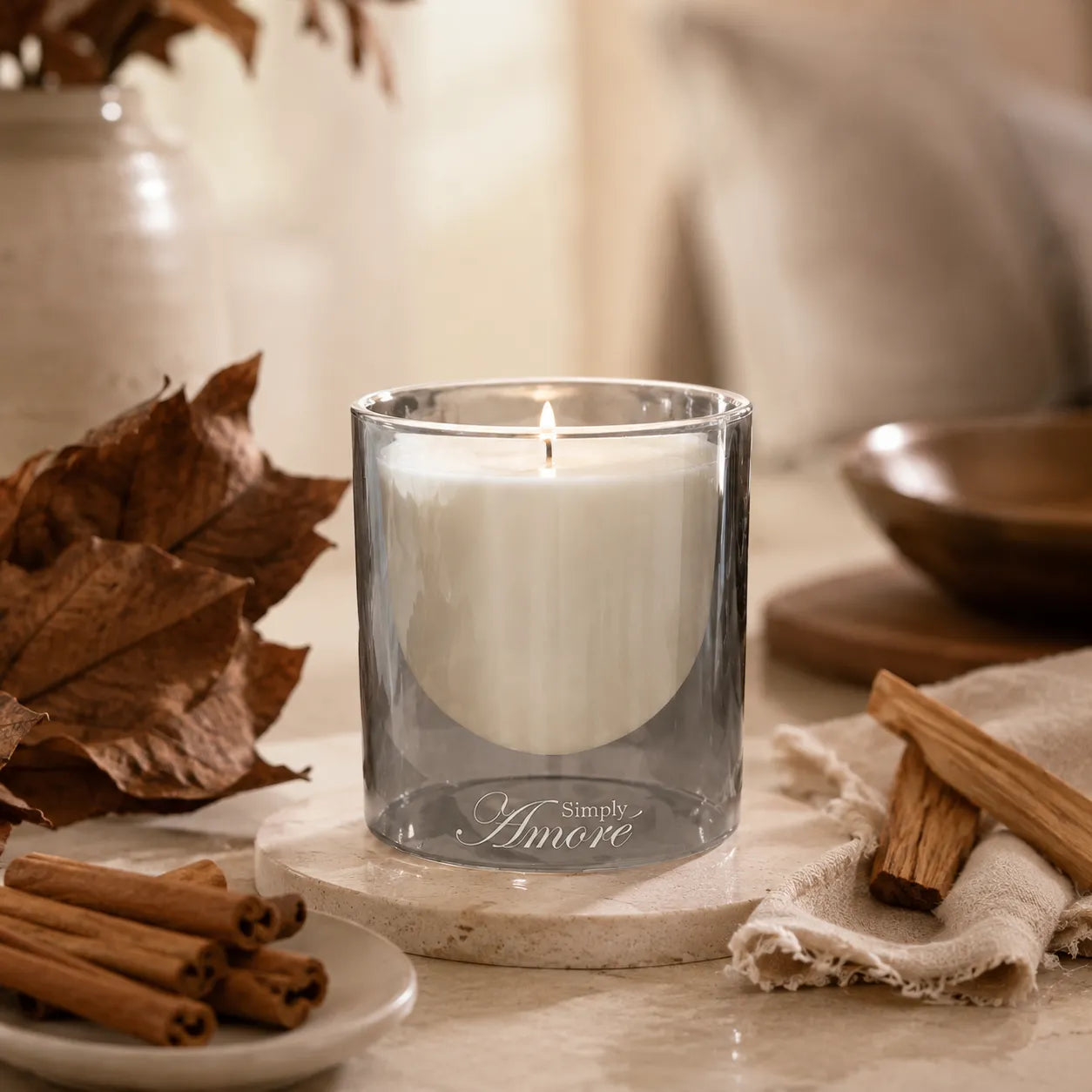 A lit white candle in a clear smoky glass holder sits on a stone coaster on a tabletop, styled with dried leaves, cinnamon sticks, and a neutral fabric in a warm indoor setting.