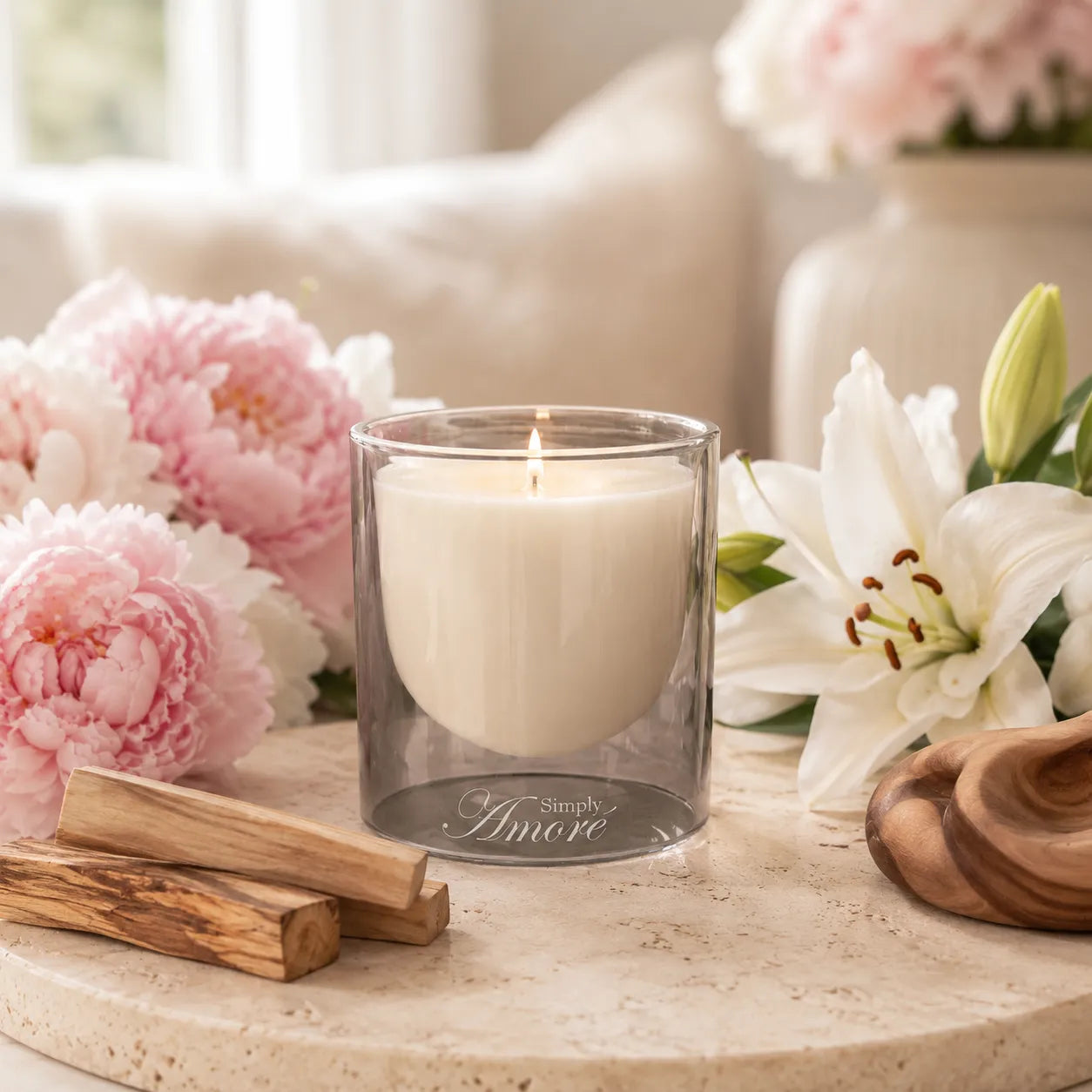 A lit scented candle in a clear glass jar sits on a stone surface surrounded by pink and white flowers, wood pieces, and a wooden bowl in a softly styled indoor setting.