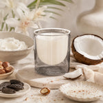 A glass jar candle with white wax sits on a stone board surrounded by coconut halves, dried dates, nuts, cream, and soft floral decor. The candle is the main product, photographed in a styled, warm setting.