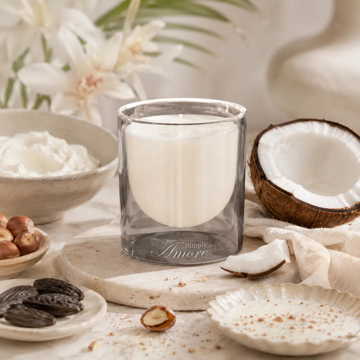 A glass jar candle with white wax sits on a stone board surrounded by coconut halves, dried dates, nuts, cream, and soft floral decor. The candle is the main product, photographed in a styled, warm setting.