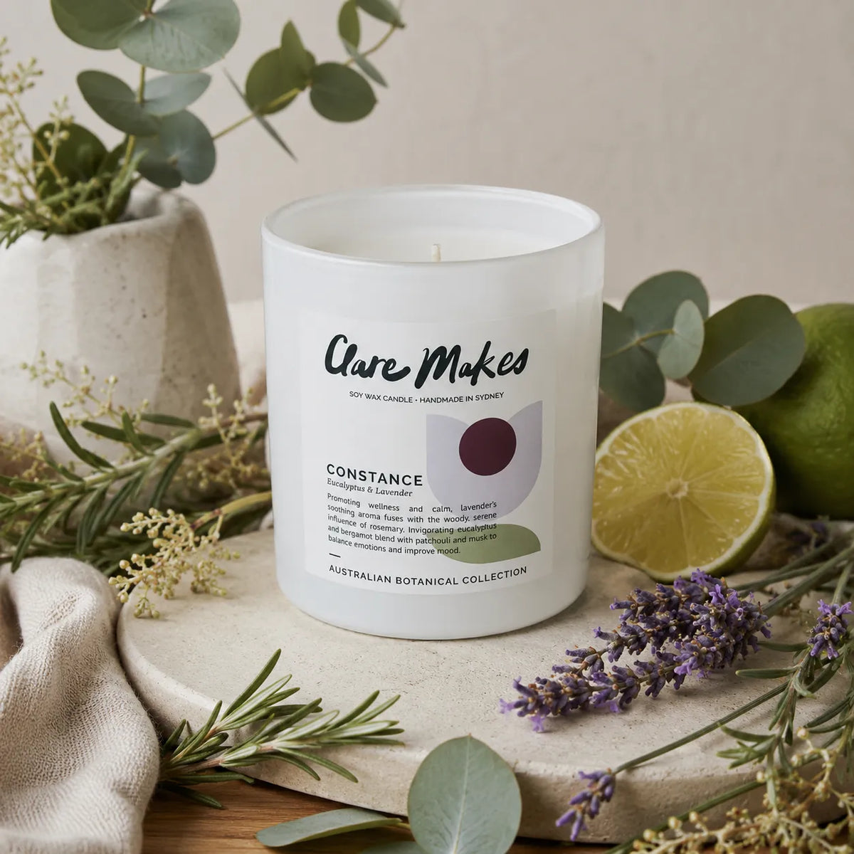 A frosted glass candle jar labeled 'Clare Makes Constance' styled on a neutral surface with eucalyptus, lavender, rosemary, a lime half, and a green lime, against a soft beige background.