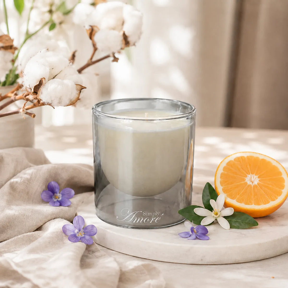 A glass jar candle with a frosted off-white wax inside, styled on a marble tray with a sliced orange, small purple and white flowers, and cotton stems in a soft, sunlit setting.