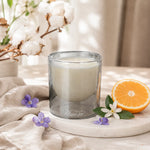 A glass jar candle with a frosted off-white wax inside, styled on a marble tray with a sliced orange, small purple and white flowers, and cotton stems in a soft, sunlit setting.