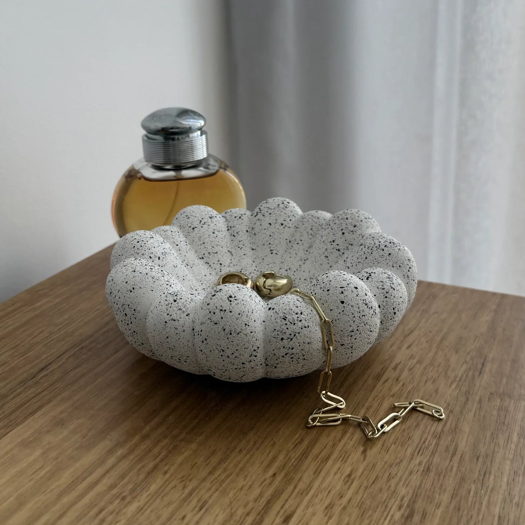A scalloped white resin clutch purse with black speckles, a gold heart clasp, and a gold chain strap, resting on a wooden table next to a perfume bottle.