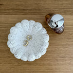Two gold hoop earrings displayed on a white scalloped speckled ceramic dish, with a small amber bottle next to it on a wooden tabletop.
