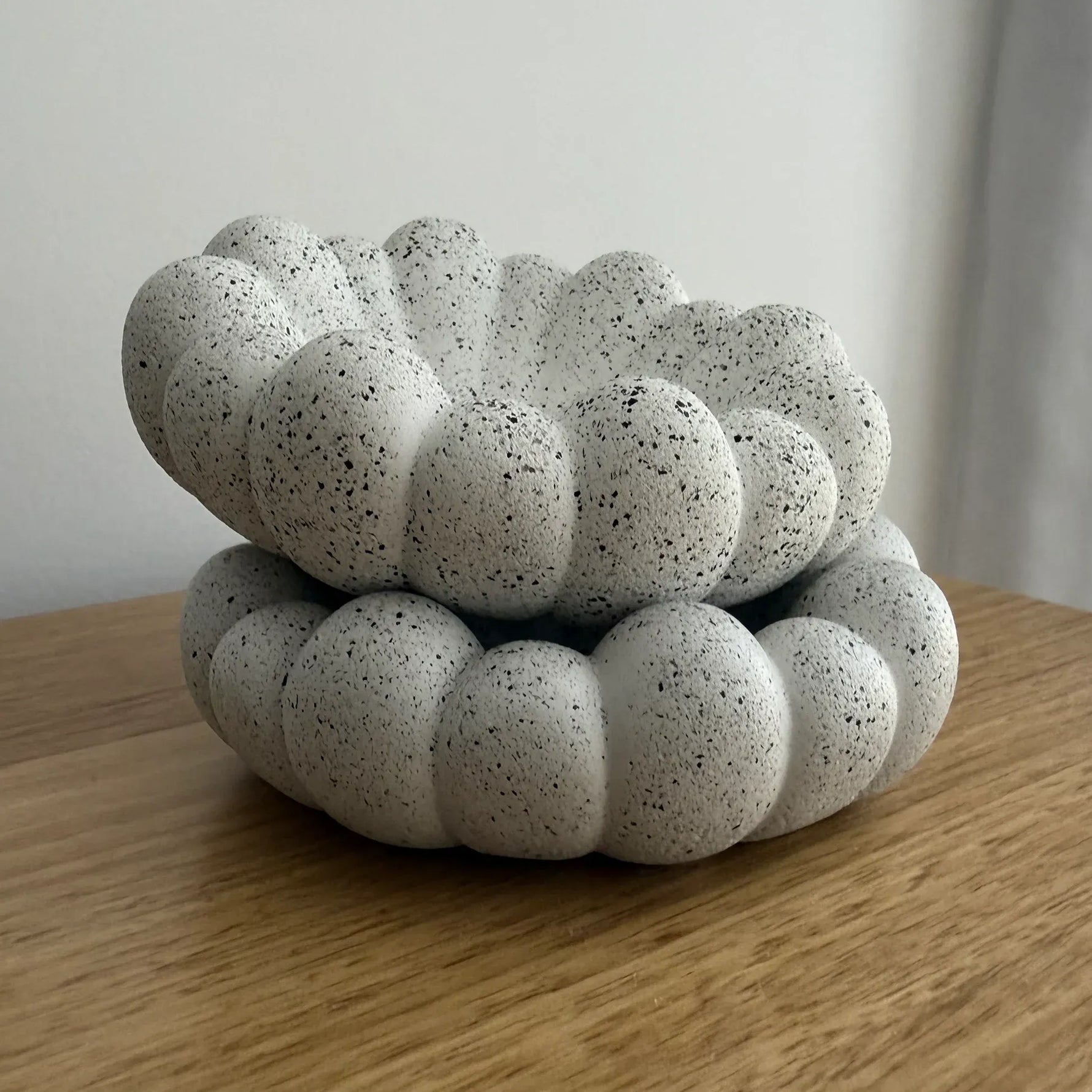 A small decorative ceramic container with a scalloped, bubble-like rim, shown stacked on top of a matching base. The ceramic is speckled off-white with dark flecks, placed on a wooden surface against a plain light background.