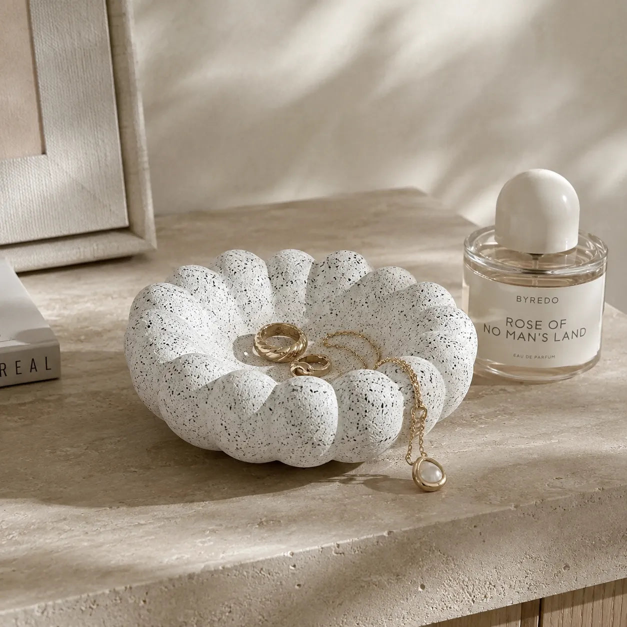 A scalloped white ceramic jewelry tray with black speckles sits on a beige stone surface, styled with gold rings and a chain necklace, next to a perfume bottle.