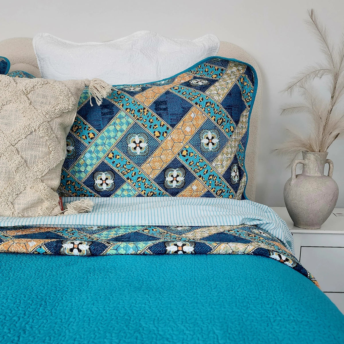 Bed with a decorative quilt featuring blue and gold patterns, white pillows, and a vase on a nightstand.