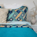Bed with a decorative quilt featuring blue and gold patterns, white pillows, and a vase on a nightstand.
