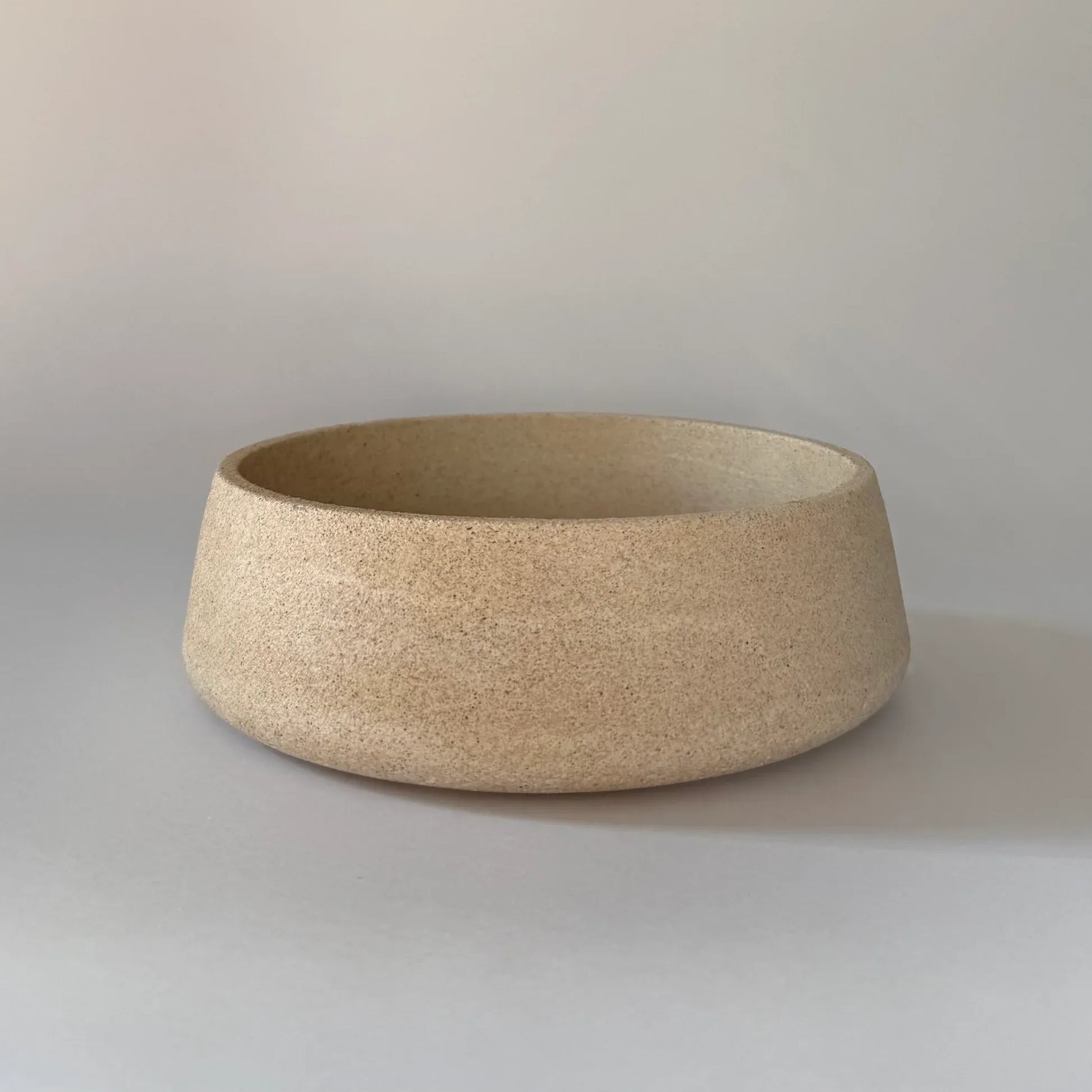 A round, shallow ceramic bowl with a textured, speckled beige finish, photographed on a neutral light gray background.