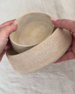 Two hands holding a small, round, speckled beige ceramic bowl against a neutral, light beige fabric background.