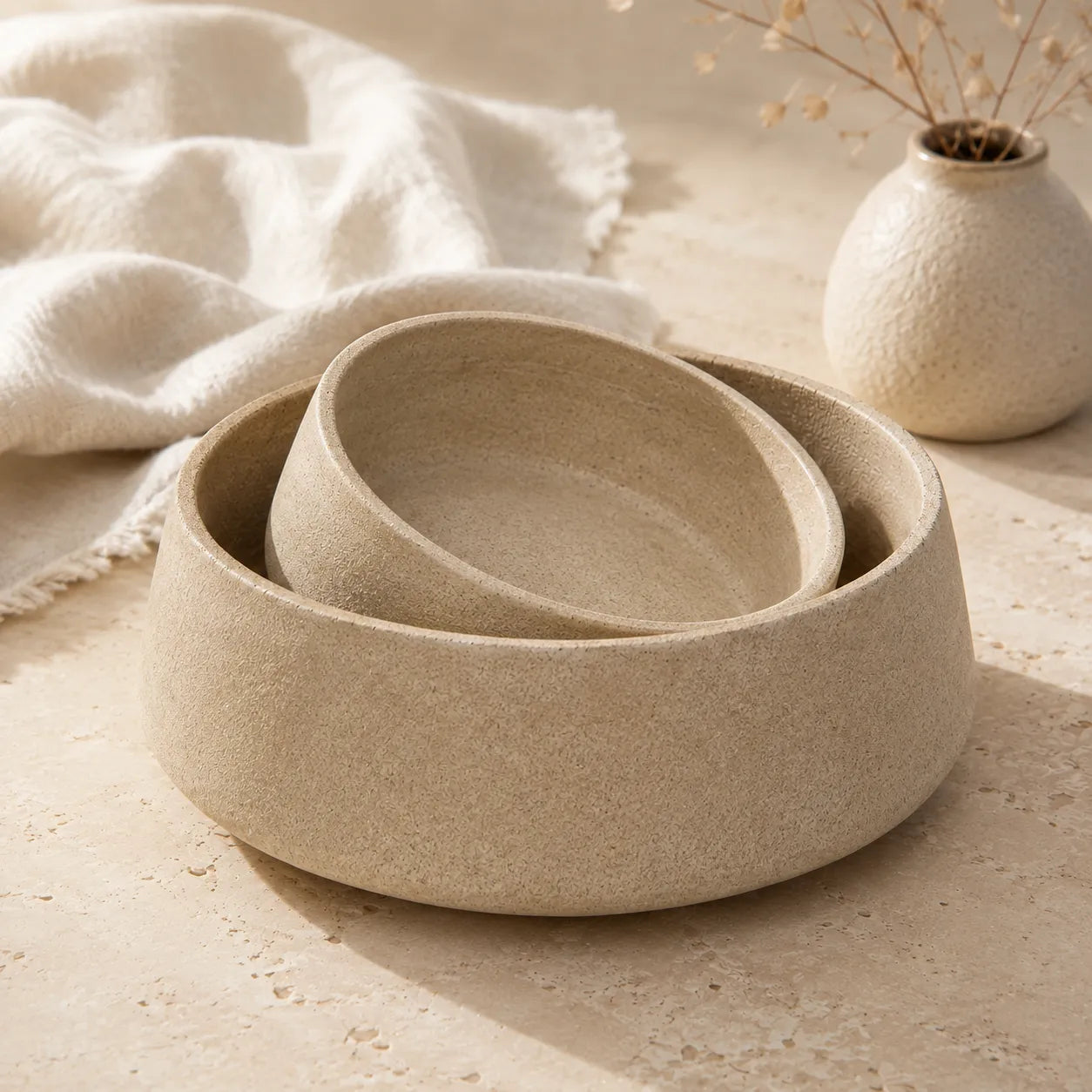 Three nested round ceramic bowls in a warm sandy beige color with a textured matte finish, styled on a stone surface with a soft white cloth and a small vase in the background.