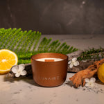 Bronze candle with 'LUNAIRE' branding on a marble surface with lemon, flowers, and greenery.